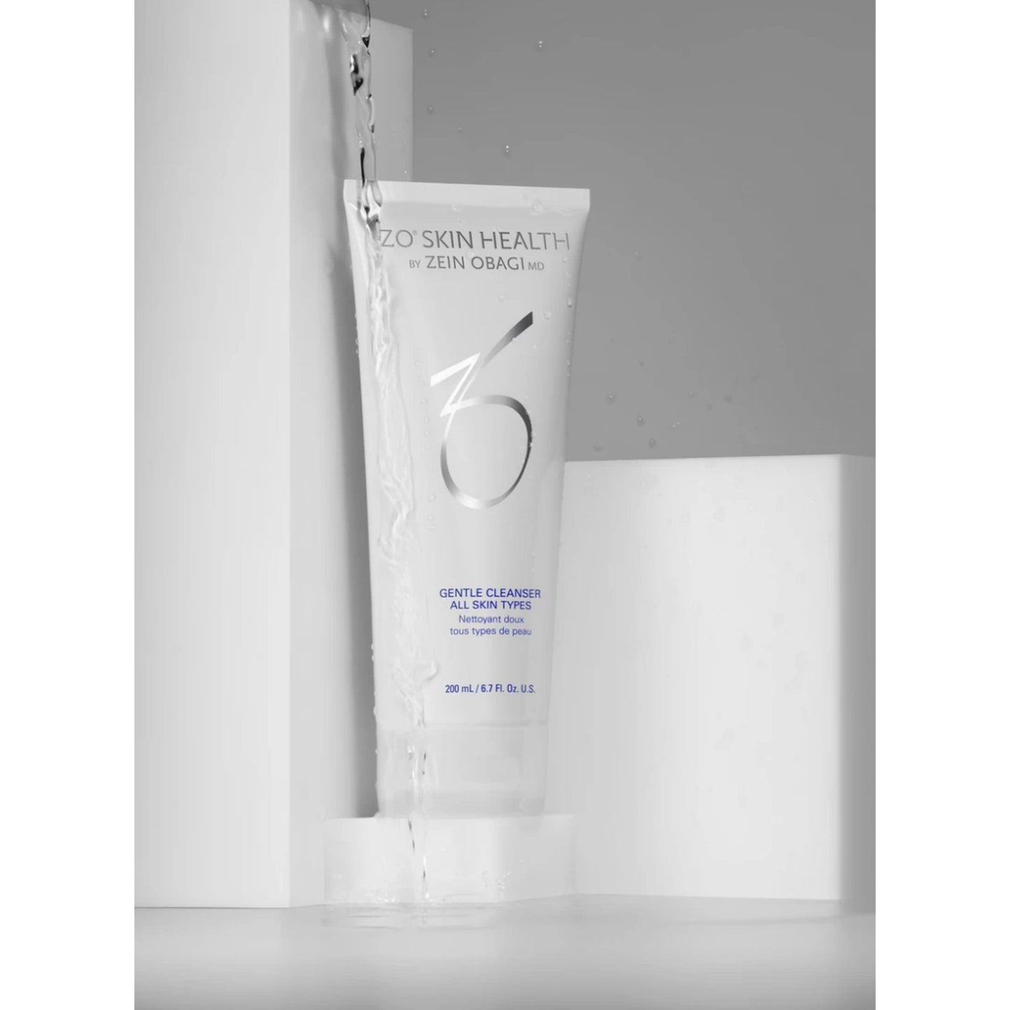 A white tube of Zeno Skin Health + Zein Obagi Gentle Cleanser All Skin Types 200mL, shown standing upright in a minimalist studio setup with water droplets splashing around it.