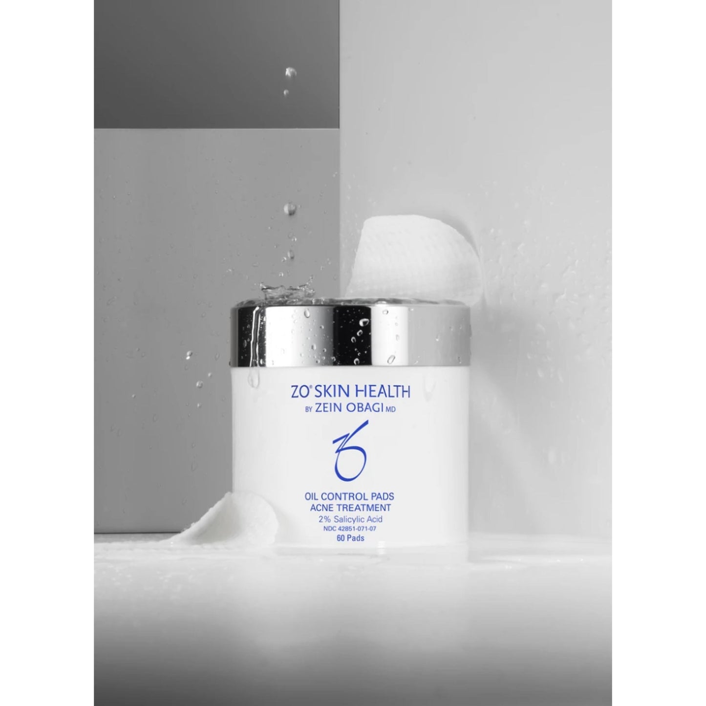 A white oil control pads jar with a reflective silver lid, labeled 'ZO'S SKIN HEALTH by ZEIN OBAGI', set against a minimalist gray background with water droplets and a cotton pad.